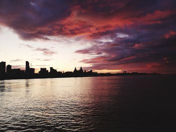 Silhouette buildings by river against sky during sunset