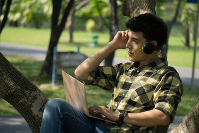 Low angle view of young man wearing headphones while laptop at park 