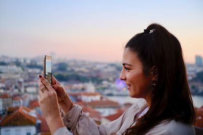 Side view of young woman photographing with mobile phone in city against clear sky 