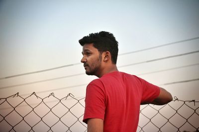 Thoughtful young man standing by chainlink fence against clear sky