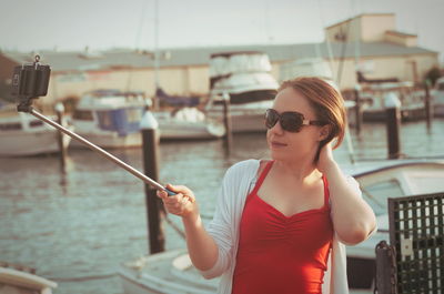 Young woman holding monopod while taking selfie through smart phone at harbor