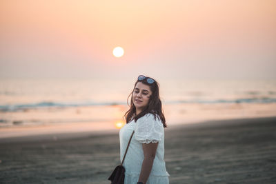 Portrait of beautiful young woman smiling and standing at beach during sunset with eyes closed 