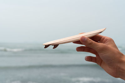 Cropped hand of person holding small surfboard against sky