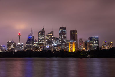 Illuminated buildings by river against sky at night