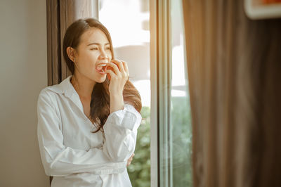 Beautiful young woman standing against window at home