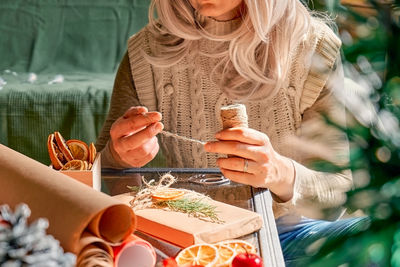 Blond woman wrapping presents in recycled card and decorated it with dried oranges and fir branches.