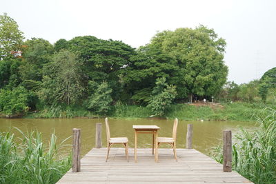 Empty chairs and table by lake against trees