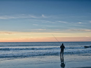 Silhouette man fishing at beach against sky during sunset