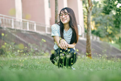 Cheerful young woman crouching on grass against building at park