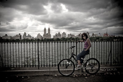 Woman on bicycle by railing and river against cloudy sky at central park