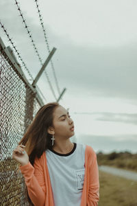 Young woman looking away while standing by fence against cloudy sky