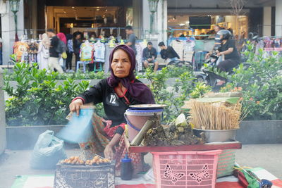 Portrait of woman preparing food while sitting against plants in city