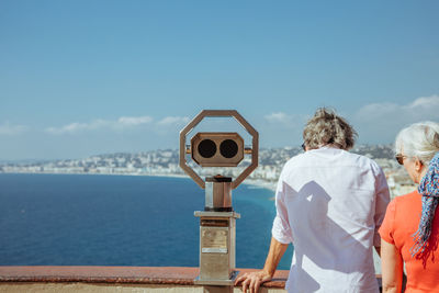 Rear view of man and woman standing by coin-operated binoculars against sky