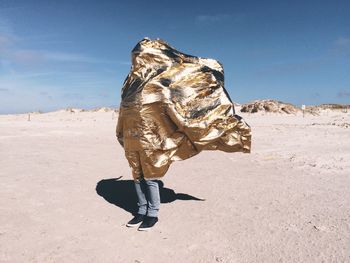 Person covered with golden sheet standing at sankt peter-ording beach against sky