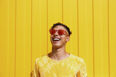 Happy young man wearing red sunglasses in front of yellow wall