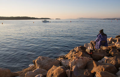 Rear view of man sitting on rock by sea against sky