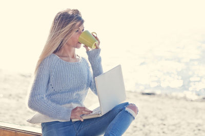 Beautiful woman drinking coffee while using laptop at beach