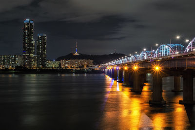 Illuminated bridge over river by buildings against sky at night