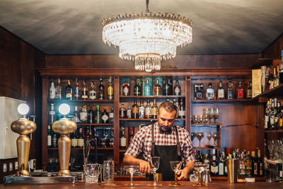 Crop brutal male bartender in apron pouring drink from bottle in glass placed on counter with bottles of alcoholic drinks