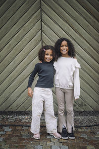 Portrait of smiling female siblings standing with arms around in front of wall