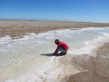 Boy crouching on frozen beach against clear blue sky