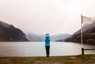 Rear view of woman photographing with digital tablet by lake during foggy weather
