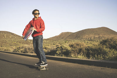 Smiling teenage boy skateboarding on road against clear sky