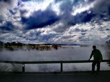 Rear view of man standing by lake against cloudy sky