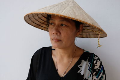 Mature woman wearing asian style conical hat while looking away against white wall