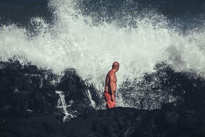 Shirtless man standing on rock while looking at splashing wave