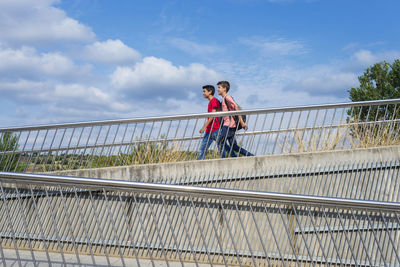 Side view of friends with backpacks walking on footbridge against blue sky during sunny day