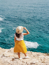Young woman in yellow skirt standing on edge of cliff above sea.