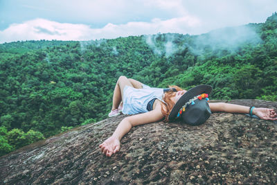 Young woman wearing hat lying on rock against sky