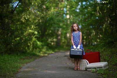 Full length of cute girl with luggage standing on road in forest