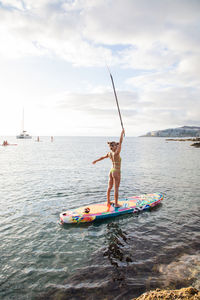 Full body side view of unrecognizable slim active female surfer in swimwear holding paddle in raised hand while standing on paddle board floating in sea water near coast