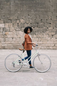 Side view of black woman in smart casual style walking along street with bike and looking at camera