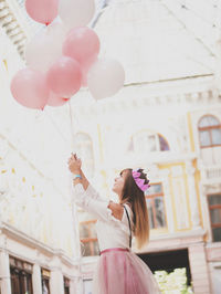 Side view of young woman holding pink balloons while standing against building