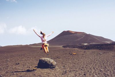 Full length of woman jumping over landscape with mt etna in background against sky