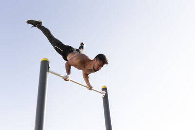 Active male athlete exercising on horizontal bar in park against clear sky
