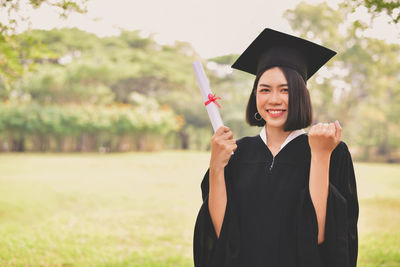 Portrait of young woman in graduation gown holding certificate while standing at park