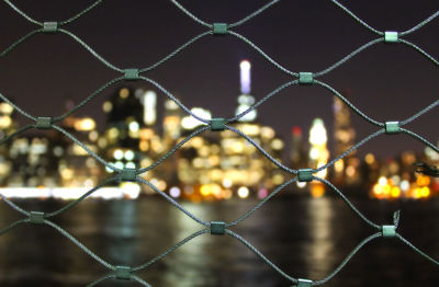 Illuminated cityscape seen through chainlink fence against sky at night