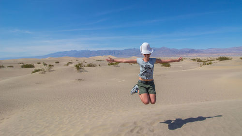 Full length of woman with arms outstretched jumping at desert against sky