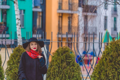 Portrait of woman wearing hat standing against fence in city