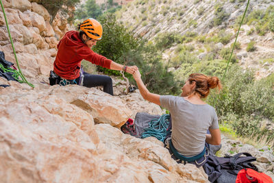 From above back view of sportive young female alpinists bumping fists together while resting on cliff in sunny day