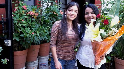 Portrait of smiling women with bouquet standing against potted plants