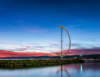 Illuminated street lights by lake against sky at sunset