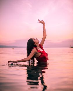 Woman with arms raised in sea against sky during sunset