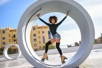Full length of young woman with roller skates standing in circle structure against sky