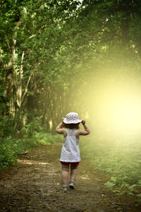 Rear view of girl wearing hat walking on field against trees in forest