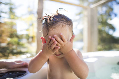 Shirtless wet baby boy covering eyes with hands while swimming in pool